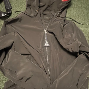 Svart vindjacka från Moncler - Säljer en svart vindjacka från Moncler med huva och dragkedja framtill. Jackan har Moncler-logga på bröstet och en snygg, minimalistisk design. Perfekt för dig som vill ha en stilren jacka med exklusiv känsla.