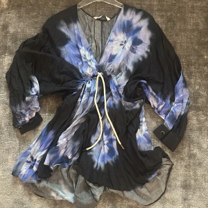 Blå och svart omlottklänning med knytband - Säljer en flowy kortklänning i blått och svart batikmönster med v-ringning och knytband i midjan. Klänningen har långa, vida ärmar med manschett och knappdetalj. Perfekt för dig som gillar unika mönster och en avslappnad stil. Zara 