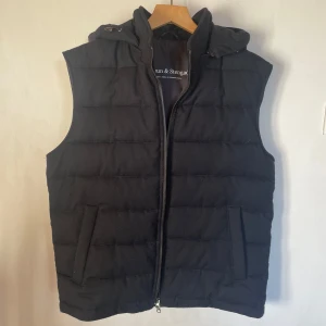 Marinblå dunväst från Bruun & Stengade i storlek M - Snygg marinblå dunväst från bruun & stengade med huva och dragkedja framtill i storlek M som är insperad av de trendiga västarna från stenströms. Västen har två fickor på sidorna quiltad design som ger en modern look. Perfekt att bära nu på sommarkvällarna. Nypriset på denna västen ligger på 2200kr och har använts 1-3 gånger!! Skriv vid funderingar!!