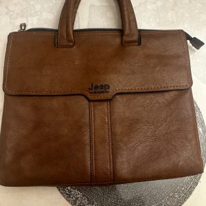 Brun portfölj från Jeep - Snygg brun portfölj från Jeep med två handtag och dragkedja upptill. Väskan är gjord i skinnimitation och har flera fack inuti för enkel organisering. Perfekt för att bära laptop, papper och andra viktiga grejer.
