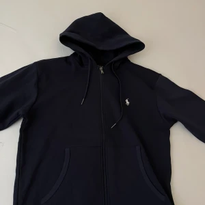 Mörkblå hoodie från Polo Ralph Lauren - Säljer en stilren mörkblå hoodie från Polo Ralph Lauren med dragkedja och huva. Tröjan har två fickor framtill och den klassiska broderade loggan på bröstet. Perfekt för en avslappnad och snygg look.