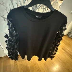 Svart tröja med 3D-blommor på ärmarna - Säljer en svart långärmad tröja unika 3D-blommor längs hela ärmarna. Tröjan har rund halsringning och en rak passform. Perfekt för dig som vill sticka ut med en cool detalj på ärmarna. Tröjan är lite noppig. 