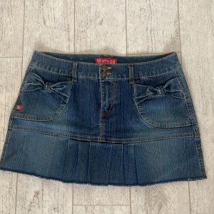 Mini Jeanskjol med rosetter  - Säljer en blå jeanskjol från Plugg med två söta rosetter på fickorna framtill. Kjolen har klassiska jeanssömmar och är i kort modell. Midjemått 40 rakt över och längd 32 cm
