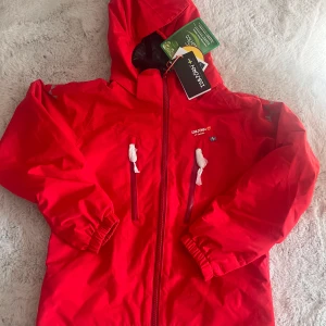 Röd vindjacka från Isbjörn of Sweden - Snygg röd vindjacka med huva från Isbjörn of Sweden. Jackan har dragkedja framtill, två bröstfickor med vita dragkedjor och elastiska muddar vid ärmsluten. Perfekt för blåsiga dagar och utomhusaktiviteter. Hög vattenpelare och bästa jackan för barn om du frågar mig. Vi hade två så den här blev inte använd och nu är den förliten. Storlek 122/128