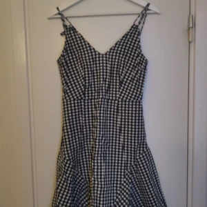 Svartvit rutig klänning med smala axelband - Säljer en svartvit rutig klänning med smala axelband och figurnära passform. Klänningen har en klassisk v-ringning och är perfekt för varma dagar. Mönstret är tidlöst och passar till många olika stilar.