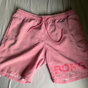 Rosa badshorts från Björn Borg - Säljer ett par snygga rosa badshorts från Björn Borg med snörning i midjan och tryckt logga på ena benet. Shortsen har elastisk midja, sidofickor och en bakficka. Perfekta för stranden eller poolen! Obs ett av snörena har tappat sin grejs.