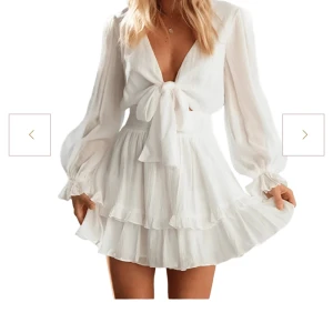 Vit volangklänning med rosett - Superfin vit kortklänning med volanger och långärmade puffärmar. Djup v-ringning och knyt framtill ger en romantisk vibe. Tillverkad i ett lätt och luftigt material. Perfekt för sommarens alla dagar och inte minst studenten!! Köp och få den skickad samma dag💛OBS: lånade bilder från hemsidan! Klänningen är inte använd utan endast testad.