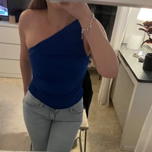 Blå one shoulder topp - Säljer en snygg blå one shoulder topp med tight passform. Toppen är ärmlös och har en asymmetrisk design som ger en trendig look. Perfekt att styla med jeans eller kjol för en cool outfit.