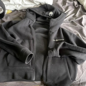 Svart hoodie från Balracing - Svart hoodie från Balracing med dragkedja och huva. Mjuk insida och två fickor framtill. Logga på bröstet och ett tryck på huvan. Perfekt för en avslappnad stil.