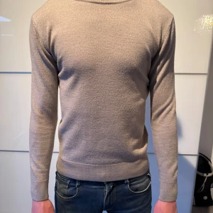 Beige stickad tröja - Säljer en stilren beige stickad tröja med rund halsringning och långa ärmar. Tröjan har ribbade muddar vid ärmslut och nederkant, vilket ger en snygg passform. Perfekt för lager-på-lager och enkel att matcha med jeans eller chinos.