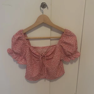 Rosa blommig croppad topp från SHEIN - Superfin rosa croppad topp från SHEIN med vita blommor. Toppen har puffiga korta ärmar med volang och knytning framtill. Perfekt för sommardagar och ger en söt och trendig look.