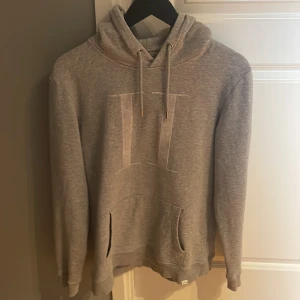 Grå hoodie från Les Deux - Säljer en stilren grå hoodie från Les Deux med huva och dragsko. Tröjan har en stor ficka framtill och diskret logga på bröstet. Perfekt för en avslappnad och trendig look.