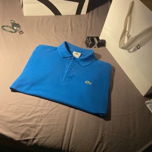 Blå pikétröja från Lacoste - Säljer en klassisk blå pikétröja från Lacoste med den ikoniska krokodilloggan på bröstet. Tröjan har krage och knäppning framtill. Perfekt för en stilren och sportig look.