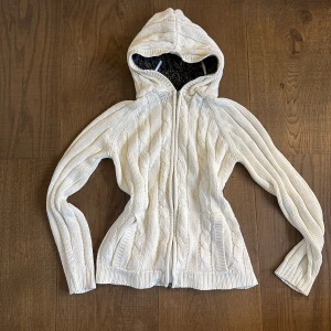 Vit stickad hoodie med dragkedja - Säljer en vit stickad hoodie med dragkedja framtill och huva. Tröjan har ett klassiskt kabelstickat mönster och ribbade muddar vid ärmslut och nederkant. Perfekt för lager-på-lager och mysiga dagar.