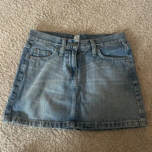 Lågmidjad jeanskjol  - Säljer en klassisk blå jeanskjol i femficksmodell med knapp och dragkedja framtill. Kjolen har bälteshällor och två bakfickor. Perfekt för dig som gillar en enkel och tidlös stil. Fint skick förutom en fläck bak!