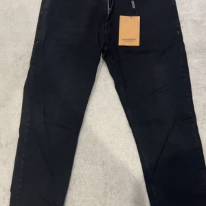 BURBERRY Jeans W38 L 35 - Säljer ett par svarta jeansbyxor från Burberry med klassisk femficksdesign och Burberry-logga på bakfickan. Byxorna har dragkedja och knapp framtill samt snygga detaljer vid midjan. Perfekta för dig som gillar stilrena och exklusiva plagg.