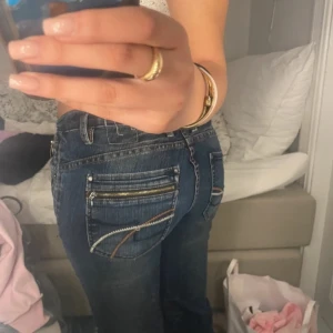 Mörkblå jeans med dragkedjedetaljer - Säljer ett par mörkblå jeans med snygga dragkedjor och kontrastsömmar på bakfickorna. Jeansen har normal passform och klassisk femficksdesign. Perfekta till vardags för en avslappnad men ändå trendig look.