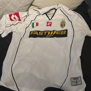 Juventus fotbollströja 02-03 - Vit vintage Juventus fotbollströja 2002-03 säsong med svarta detaljer och broderad klubbmärke på bröstet. Sponsorerna Fastweb och Lotto-logga syns tydligt. Klassisk design med svart V-ringning och italiensk flagga på bröstet. Perfekt för fans av italiensk fotboll och ett sjukt bra samlarobjekt då de inte går att hitta . Säljs annars runt 3000 kr, öppen för förslag