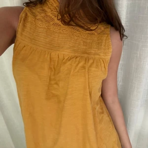 Gul ärmlös blus med spetsdetaljer - Säljer en söt orange/gul blus med spets upptill. Blusen är ärmlös och har två knappar i nacken för stängning. 