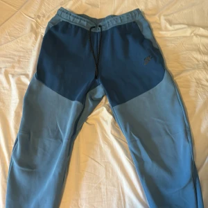 Nike blå mjukisbyxor med snörning - Säljer ett par blå Nike tech byxor från Nike i två nyanser av blått. Byxorna har snörning i midjan och en dragkedjeförsedd ficka på baksidan. Perfekta för chill eller träning.