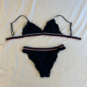 Mörkblå bikini med volang från H&M - Säljer en mörkblå bikini från H&M med volangdetaljer och smala axelband. Både överdel och underdel har en randig resår i vitt, rött och blått. Bikinin är i ett ribbat material och är i strl 36