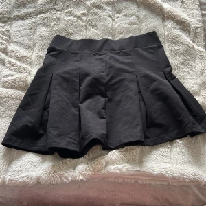 Svart kortkjol från 157 - Säljer en svart kortkjol från 157 med bred linning och utställd passform. Kjolen har diskreta veck framtill som ger extra volym och en stilren look. Perfekt att matcha med en enkel topp eller hoodie.