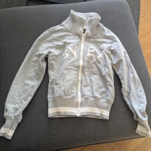 Ljusgrå zip-jacka från Svea - Ljusgrå jacka från Svea med dragkedja framtill och hög ribbad krage. Jackan har vita ribbade muddar vid ärmslut och nederkant samt broderad logga på bröstet. 