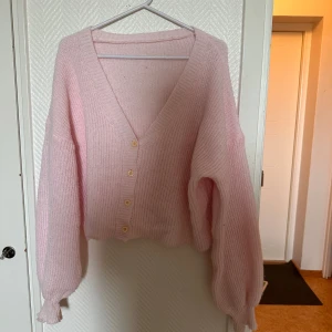 Ljusrosa stickad kofta med v-ringning - Supermjuk ljusrosa stickad kofta med v-ringning och tre små knappar framtill. Koftan har långa, lite puffiga ärmar och ribbade muddar. Perfekt för lager-på-lager och en söt touch till din outfit.
