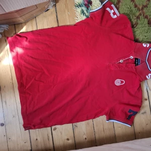 Röd pikétröja med tryck och detaljer - Röd pikétröja från La Martina med vita och blå detaljer. Tröjan har korta ärmar, nummertryck på ärmarna och broderad logga på bröstet. Klassisk krage med knappar och kontrastfärgade kanter.