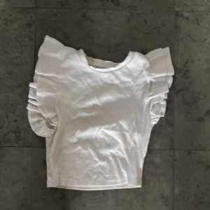 Vit ribbad topp med volangärmar - Säljer en vit ribbad topp med stora volangärmar. Toppen har rund halsringning och är tillverkad i ett mjukt material. Perfekt för dig som vill ha något enkelt men ändå med en twist.