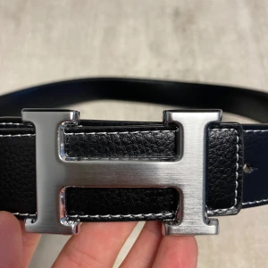 Svart skärp med H-spänne  - Säljer ett svart skärp med ett stort, silverfärgat H-spänne i metall. Skärpet är gjort i skinn med vita sömmar längs kanterna och har en klassisk, stilren design. Perfekt accessoar för att lyfta din outfit.