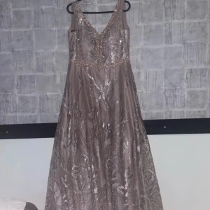 Champagnefärgad långklänning med paljetter och spetsdetaljer - Elegant långklänning i champagnefärg med v-ringning, dekorerad med paljetter och spetsdetaljer. Klänningen har smala axelband, öppen rygg med snörning och vackert glittrande mönster över hela tyget. Perfekt för bal eller fest. Säljer för 1 100kr. Kan absolut sänka priset om vi kommer överens. Har använt vid 2 tillfällen.