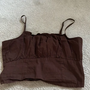 Brun croppad linnetopp från H&M Divided - Säljer en brun croppad linnetopp från H&M Divided. Toppen har smala axelband och rynkade detaljer baktill för en bekväm passform. Perfekt till varma dagar och enkel att matcha med jeans eller kjol.