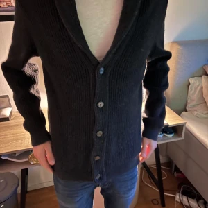 Svart ribbad kofta med knappar - Säljer en svart ribbstickad kofta med knäppning framtill. Koftan har långa ärmar och en djup v-ringning. Perfekt för lager-på-lager och enkel att matcha med olika outfits.