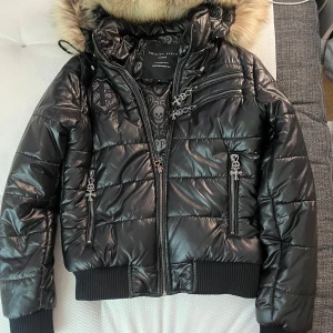Svart Y2K  pufferjacka från Philipp Plein med päls. Y2K - Säljer en svart glansig pufferjacka från Philipp Plein med päls på huvan. Jackan har flera dragkedjor, quiltad design och snyggt mönstrat innerfoder med döskallar och loggor. Perfekt för kyliga dagar.