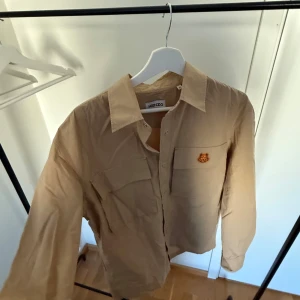 Kenzo Overshirt - Säljer en slutsåld beige overshirt från Kenzo Paris i toppskick (använd 2–3 gånger).  Lätt, luftig och stilren i sportigt 100 % bomullstyg. Med Kenzo’s ikoniska tiger broderad på bröstet och två militärinspirerade bröstfickor är den perfekt som sommarplagg för en unik och exklusiv stil.  Nypris ligger på ca 3500 kr (om du ens får tag på den) vilket gör detta plagg till sommarens kap! ☀️   Skicka DM vid intresse eller frågor. Först till kvarn! 🪭🪩✨