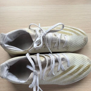 Vita Adidas fotbollsskor med guldiga detaljer - Säljer ett par vita Adidas fotbollsskor med guldiga detaljer och snörning. Skorna har en platt sula och ett mönstrat yttre i syntetmaterial. Perfekta för dig som vill sticka ut på planen!