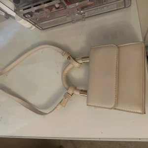 Beige mini axelväska med band - Säljer en stilren beige mini väska. Väskan har en enkel design med lock och är perfekt för att bära det viktigaste. Använt en gång💕
