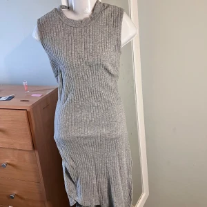 Grå ribbad ärmlös klänning - Säljer en grå ribbad klänning med ärmlös design och rund halsringning. Klänningen har en omlott-detalj nertill som ger en snygg och modern look. Perfekt för dig som gillar enkel men stilren stil.