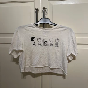 Vit croppad Peanuts t-shirt från Bershka - Säljer en vit croppad t-shirt från Bershka med tryck av snobben-karaktärer på framsidan. Perfekt till sommaren. I strl S
