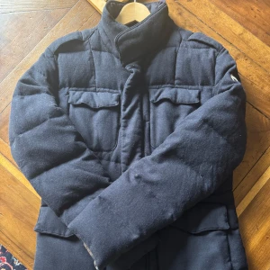 Mörkblå quiltad pufferjacka från Colmar - Säljer en mörkblå pufferjacka från Colmar med hög krage och flera fickor framtill. Jackan har quiltad design och stängs med dragkedja. Perfekt för kyliga dagar och har en stilren look. Felfri. Sitter som L/stor M