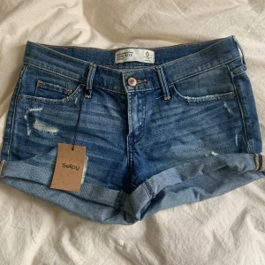 Blå jeansshorts med upprullad kant - Snygga blå jeansshorts med låg midja och upprullad kant (som går att vika ner). Klassisk femficksmodell med slitna detaljer framtill och bakfickor med dekorativa sömmar. Perfekta för varma dagar.