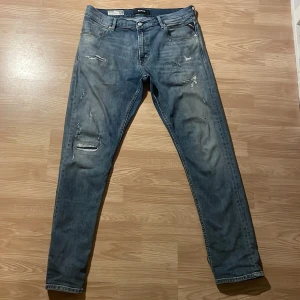 Blå jeans från Replay - Säljer ett par blå jeans från Replay med slitna detaljer och klassisk femficksdesign. Jeansen har tapered fit.