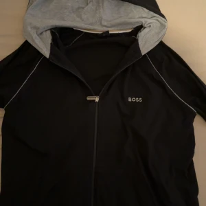 Svart hoodie med dragkedja från BOSS - Svart hoodie från BOSS med grå insida på huvan och vit piping längs axlarna. Jackan har dragkedja framtill och BOSS-logga på bröstet samt på dragkedjan. Perfekt för en avslappnad stil.