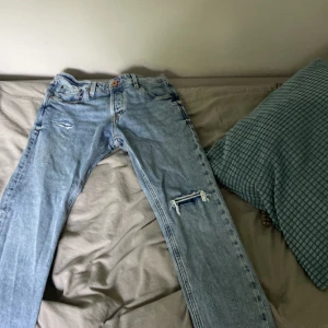 Ljusblå slitna jeans från Jack & Jones - Säljer ett par ljusblå jeans från Jack & Jones med slitningar och hål på ena benet. Klassisk femficksmodell med normal passform och knappgylf. Perfekta för en avslappnad stil.