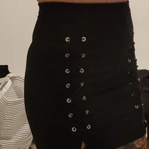 Svart kjol med snörningsdetaljer - Säljer en svart kortkjol med snygga snörningsdetaljer framtill. Kjolen har en tight passform. Passar till många olika toppar och är enkel att styla.