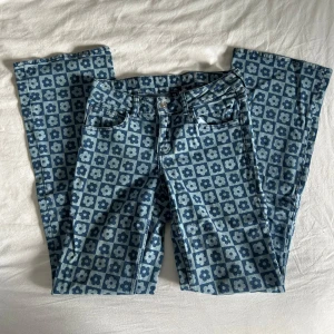 Blommiga jeansbyxor i blått från H&M - Säljer ett par unika blå jeansbyxor från H&M med blommigt rutmönster.  Byxorna är boot cut och är lågmidja, klassiska fickor fram och bak samt knapp- och dragkedjestängning och byxorna är i strl 36. Perfekta för dig som vill sticka ut med din stil!