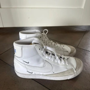 Vita Nike Blazer Mid sneakers - Säljer ett par klassiska vita Nike Blazer Mid sneakers med högt skaft och blå Nike-logga på hälen. Skorna har vit snörning, vit sula och detaljer i mocka framtill. Perfekta för en clean och sportig stil.