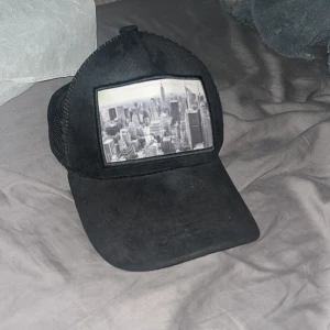 Svart keps med New York-tryck - Svart keps med mesh på sidorna och ett stort svartvitt tryck av New Yorks skyline framtill. Böjd skärm och justerbar passform baktill. Perfekt för en cool och urban stil.