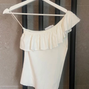 Vit oneshoulder topp med volang - Säljer en vit oneshoulder topp från Mango med volangdetalj längs axeln. Passar perfekt till sommaren
